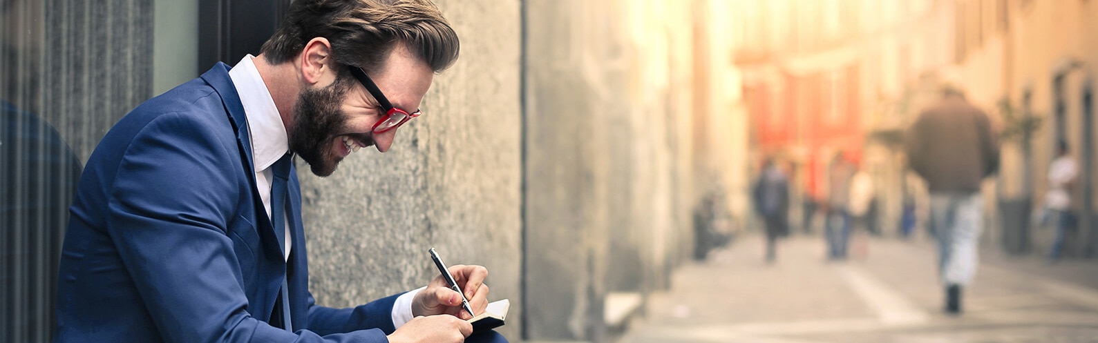 A young businessman wearing glasses sits outside, smiling and taking notes.