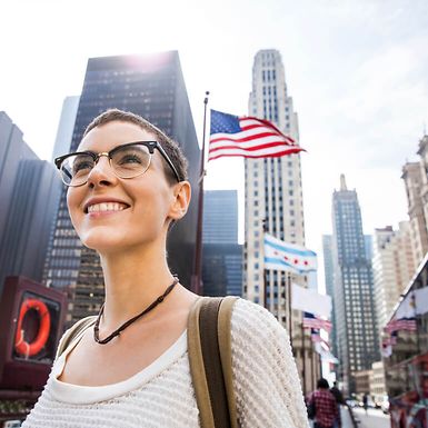 Young woman with glasses smiles in an outdoor city environment with flags in the background.