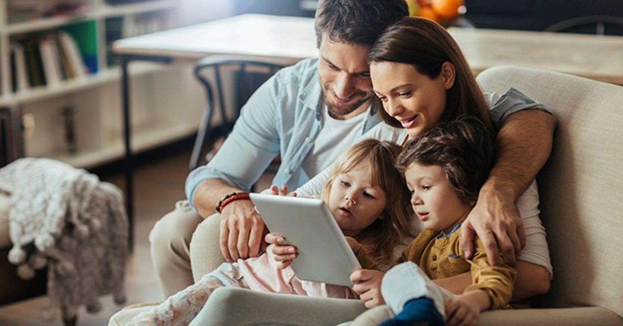 family of 4, mother, father, and two small children, sitting on couch, all looking at a tablet 